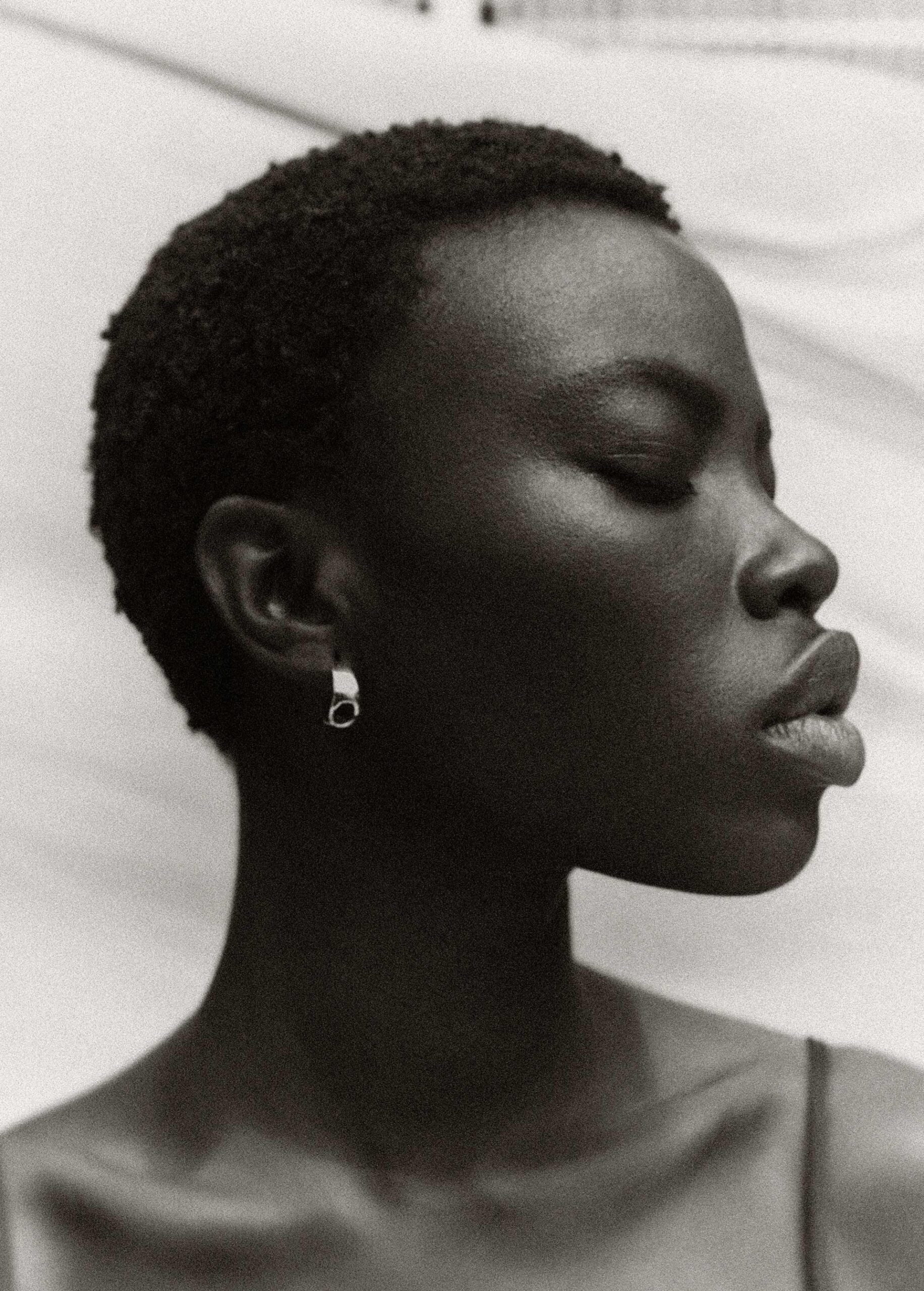 Close-up profile portrait of a black woman with short textured hair wearing a silver hoop earring against a draped white background.