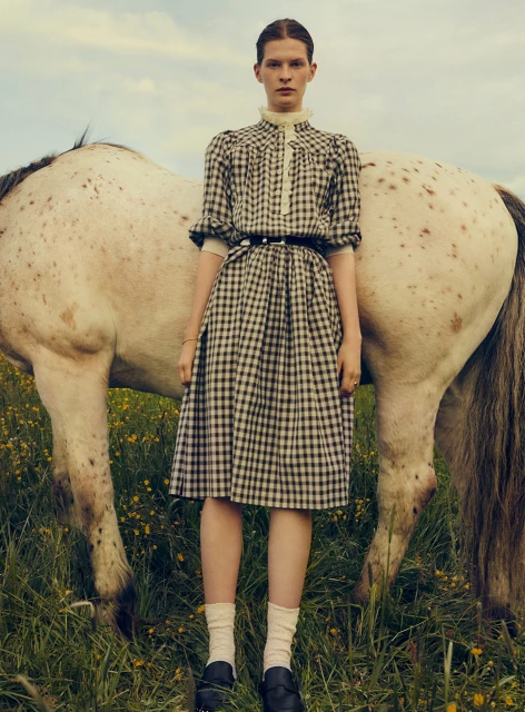 An adult female model wearing a plaid dress stands next to a white horse in an outdoor grassy field with a blue and cloudy sky.