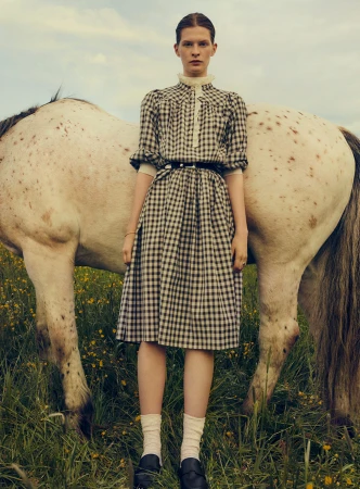 An adult female model wearing a plaid dress stands next to a white horse in an outdoor grassy field with a blue and cloudy sky.
