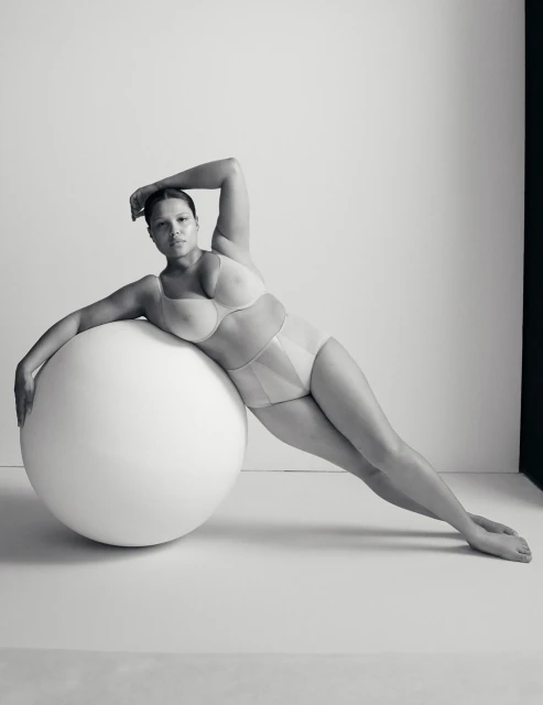 Portrait of a woman in lingerie leaning against a large white ball in a photo studio with a minimalist aesthetic and monochrome color palette.
