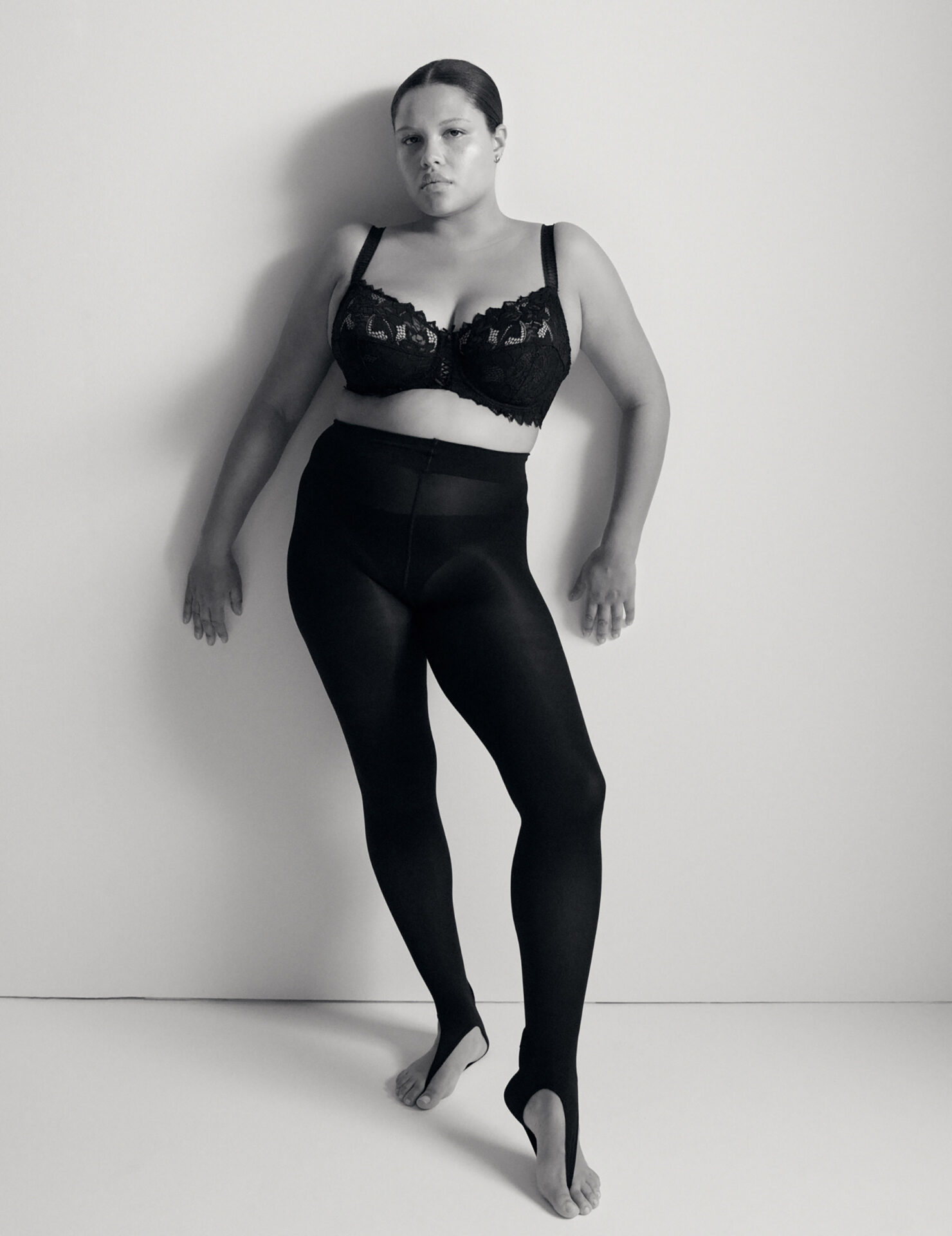 Monochrome portrait of a plus-size adult female in a black lace bra and black stirrup leggings against a light neutral background.