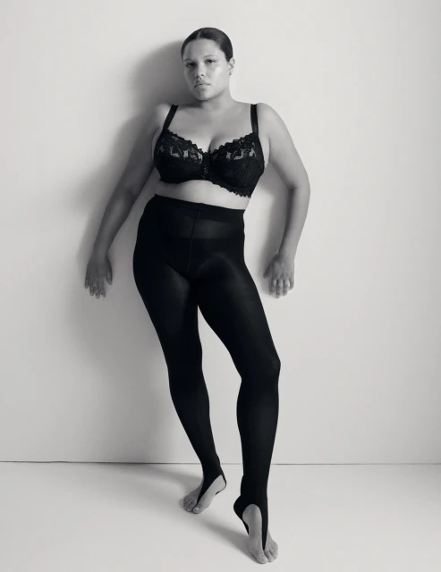 Monochrome portrait of a plus-size adult female in a black lace bra and black stirrup leggings against a light neutral background.