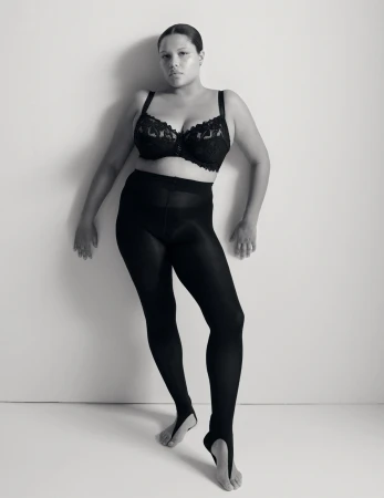 Monochrome portrait of a plus-size adult female in a black lace bra and black stirrup leggings against a light neutral background.