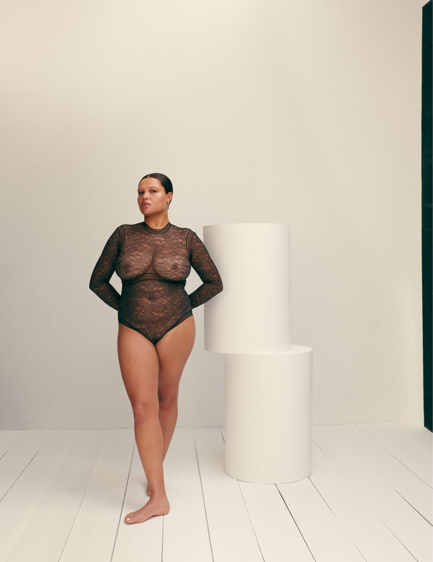 Woman wearing a black lace long sleeve bodysuit standing beside stacked white cylindrical props on a white floor