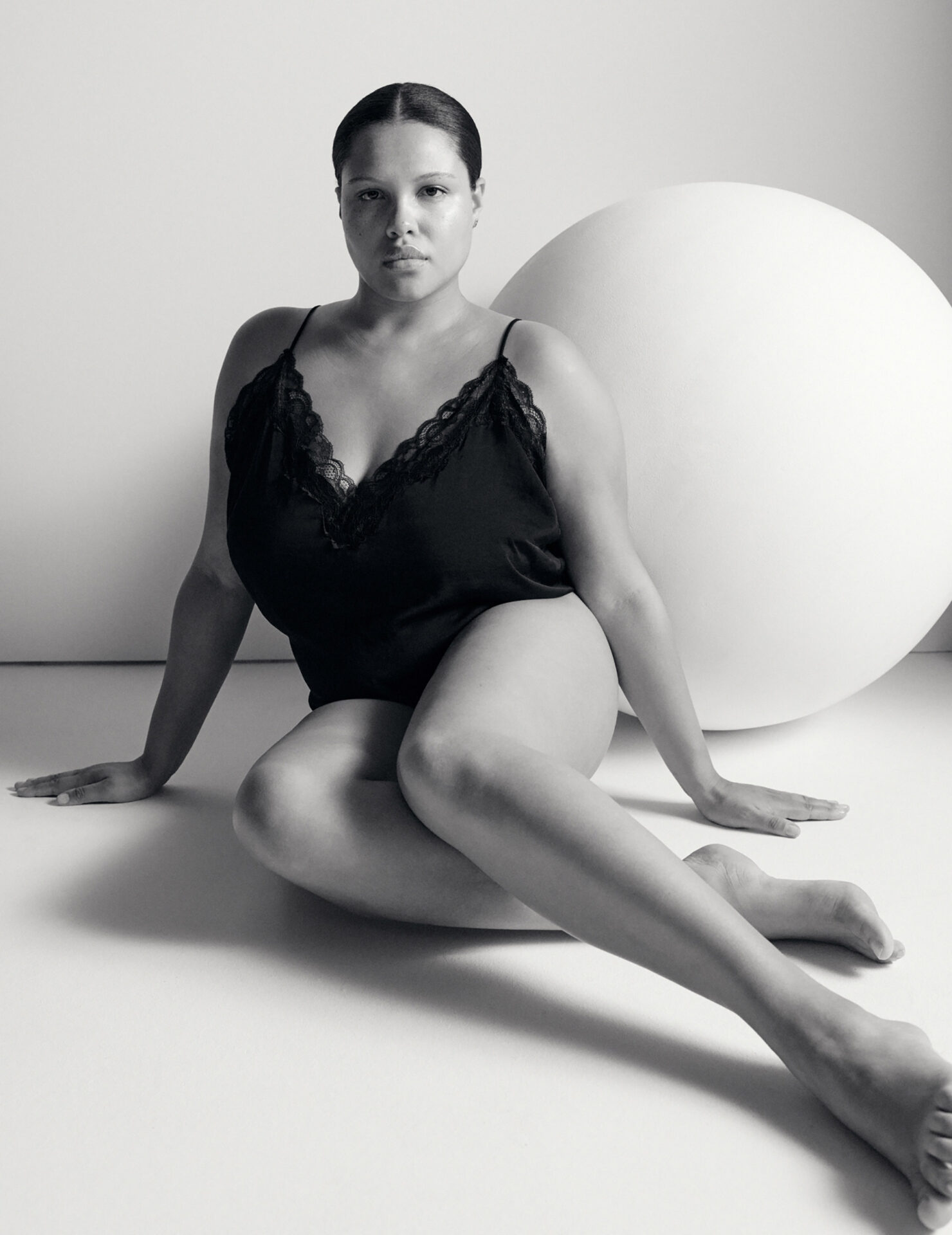 Monochrome studio portrait of a woman in a dark camisole and sitting on the floor, next to a large white sphere.
