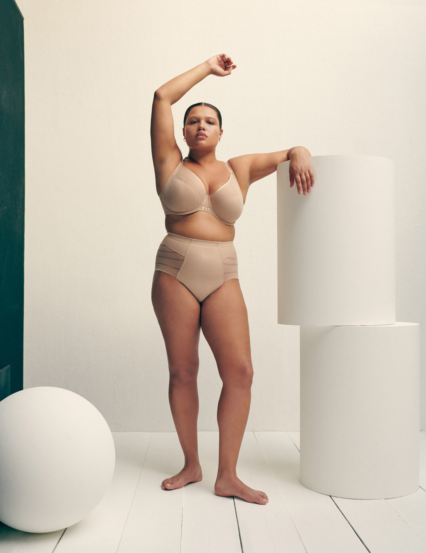 A curvy woman poses in a beige bra and high-waisted briefs in a studio with minimalist geometric props, bathed in soft, diffused light.
