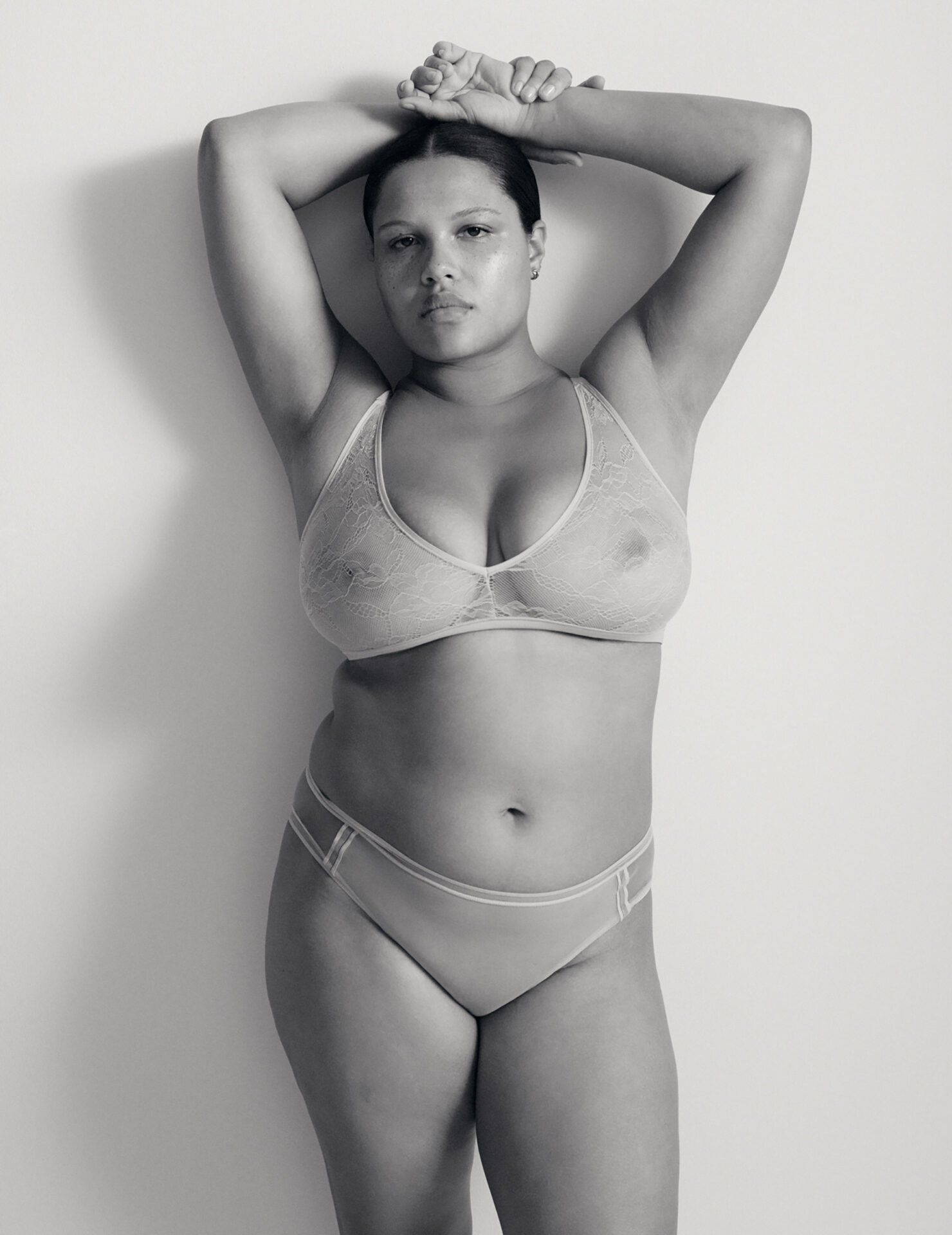 Black and white portrait of a woman wearing lingerie with her hands raised behind her head against a plain background.