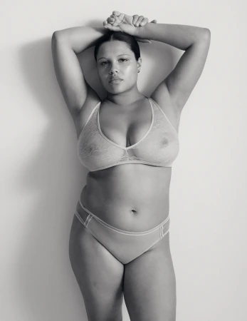 Black and white portrait of a woman wearing lingerie with her hands raised behind her head against a plain background.