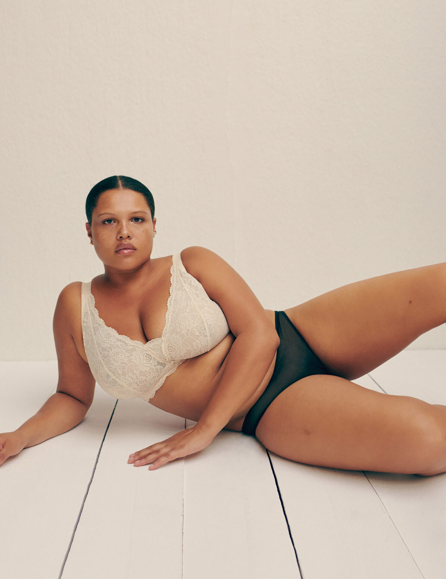 Adult woman poses in lace bra and black underwear on white wood floor in a close-up studio shot for a fashion or beauty campaign.