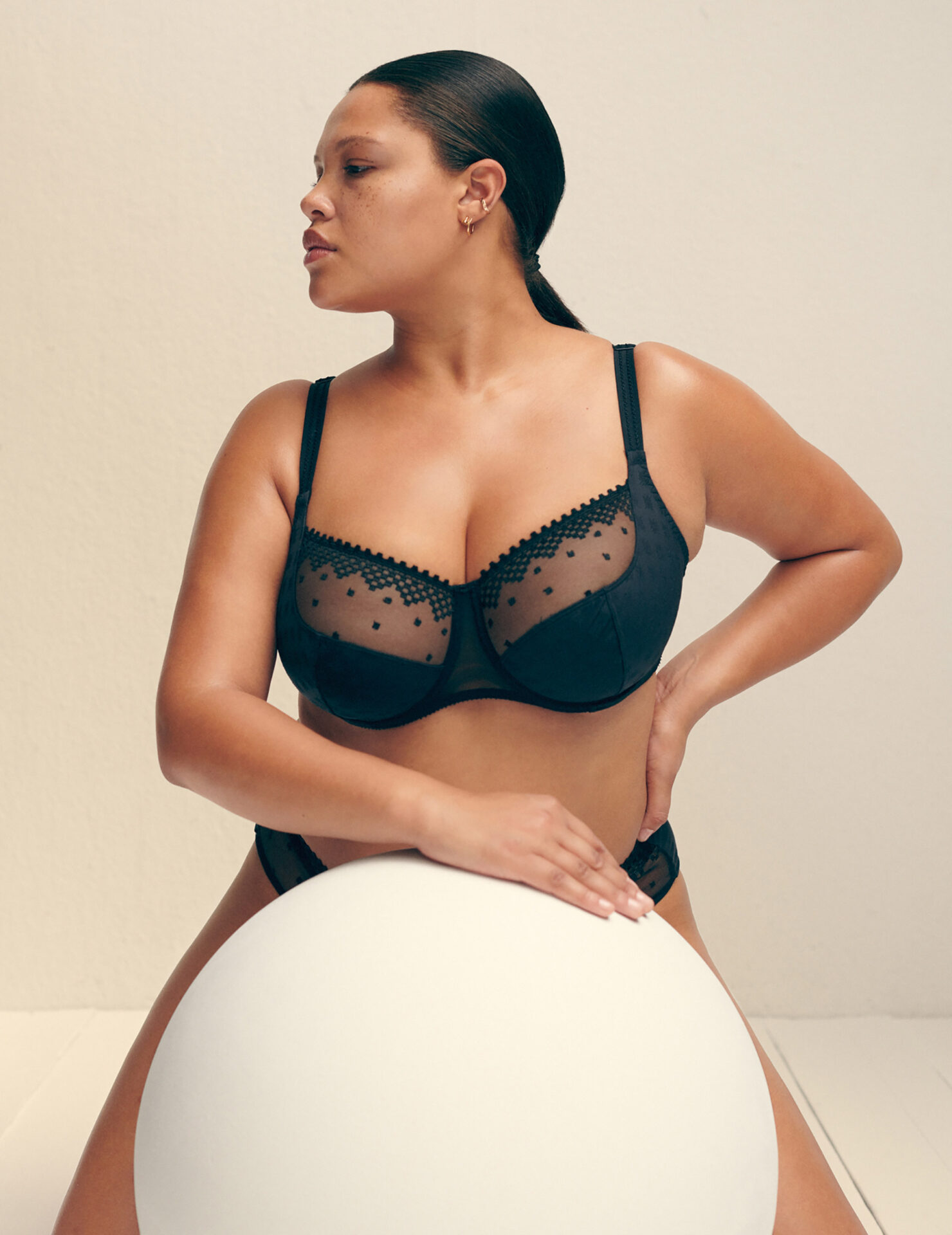 Woman in black lingerie sitting on white sphere in a brightly lit studio. Lingerie features sheer fabric and lace trim.