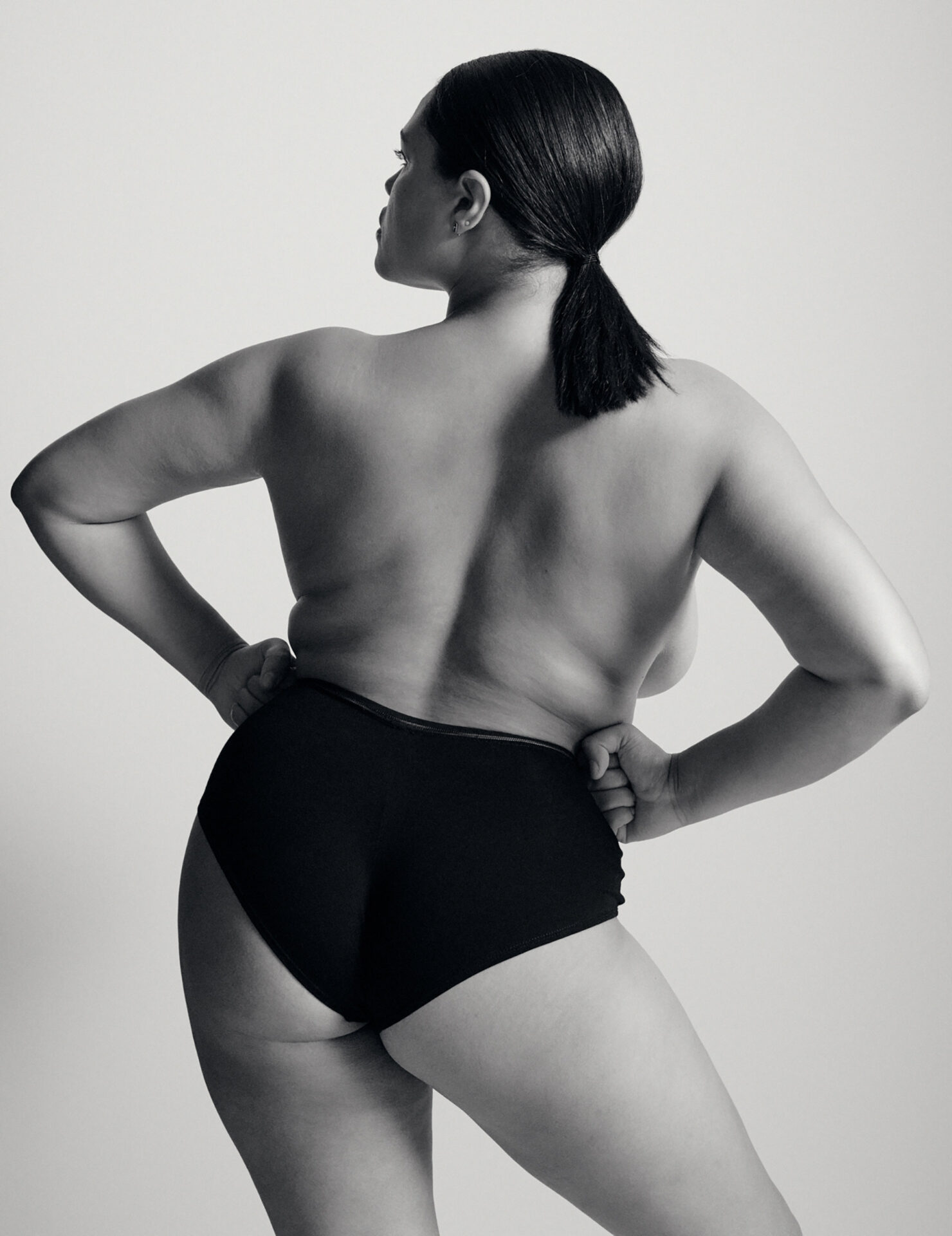 Back shot of a plus-size woman with dark hair in a ponytail, wearing dark underwear and posing in front of a plain background.
