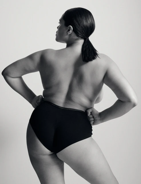 Back shot of a plus-size woman with dark hair in a ponytail, wearing dark underwear and posing in front of a plain background.