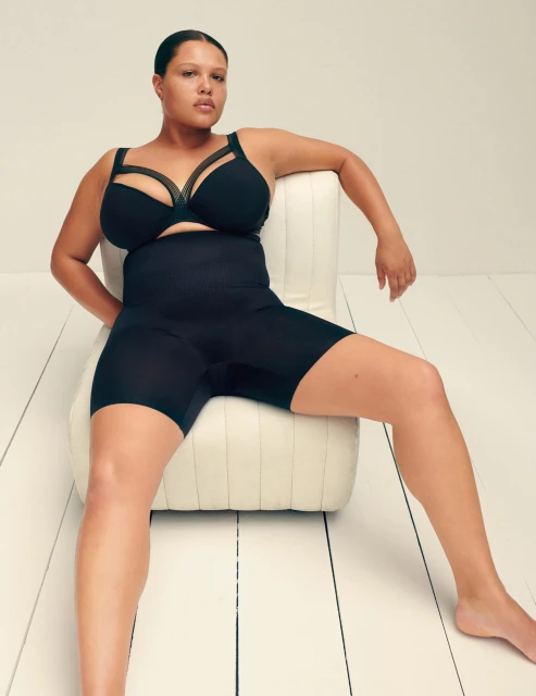 Woman in black lingerie sitting on an off-white chair in a minimalist studio setting, showcasing beauty and body positivity.