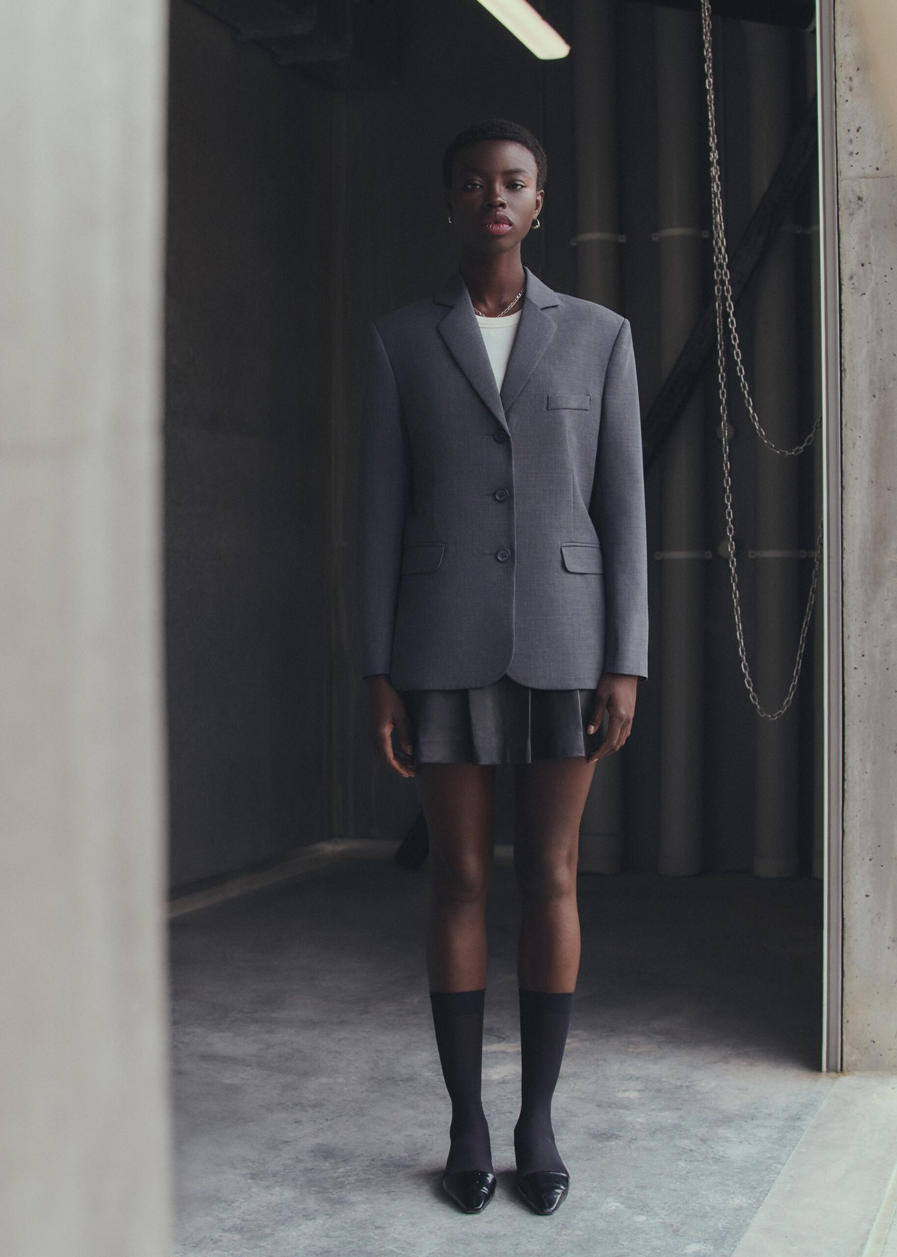 Full-length portrait of a Black female model in a gray blazer, pleated skirt, and black socks and heels in an industrial-style hallway.
