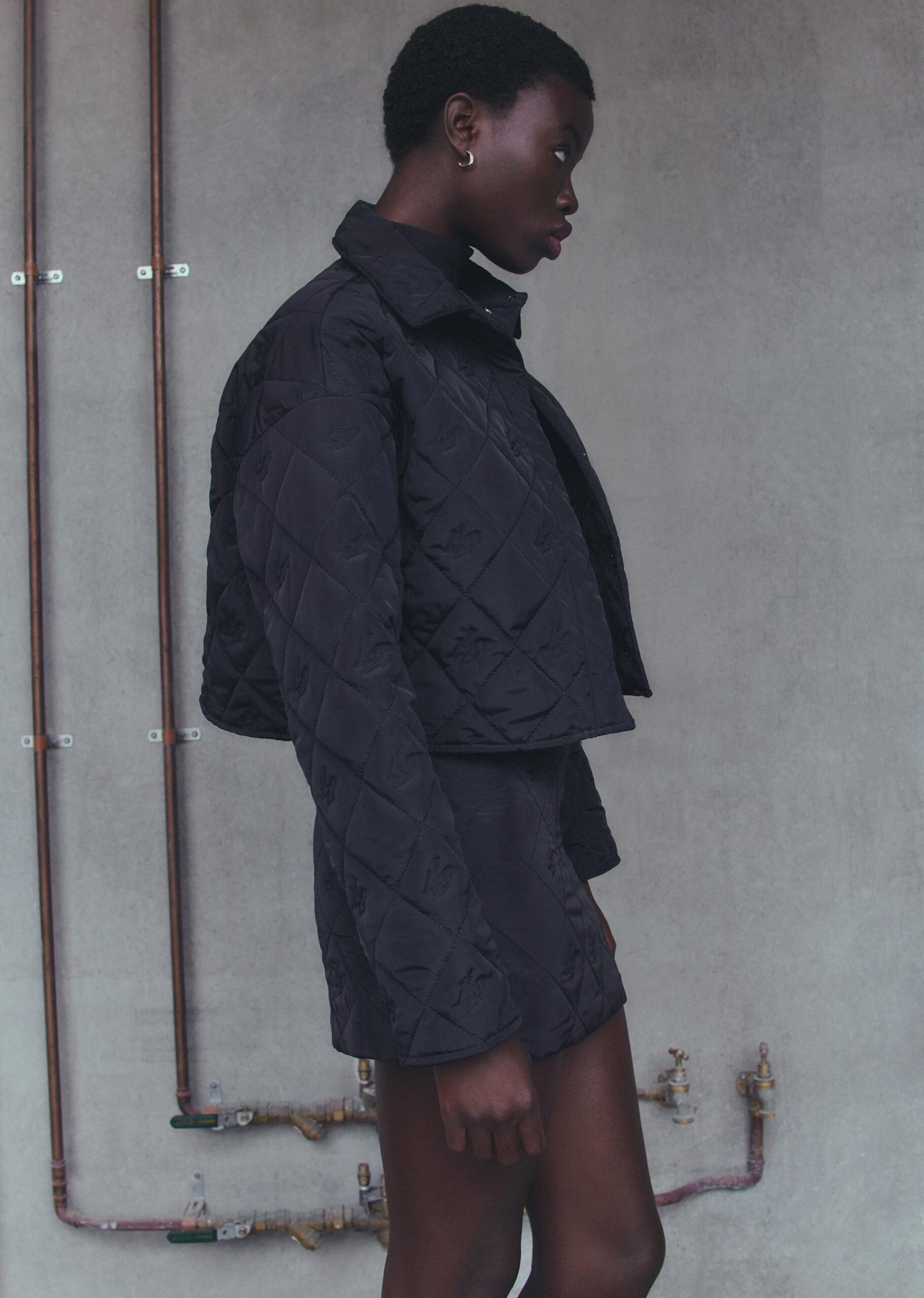 Adult female model in a short black quilted jacket and skirt poses in front of a concrete wall with copper pipes. Fashion shot.