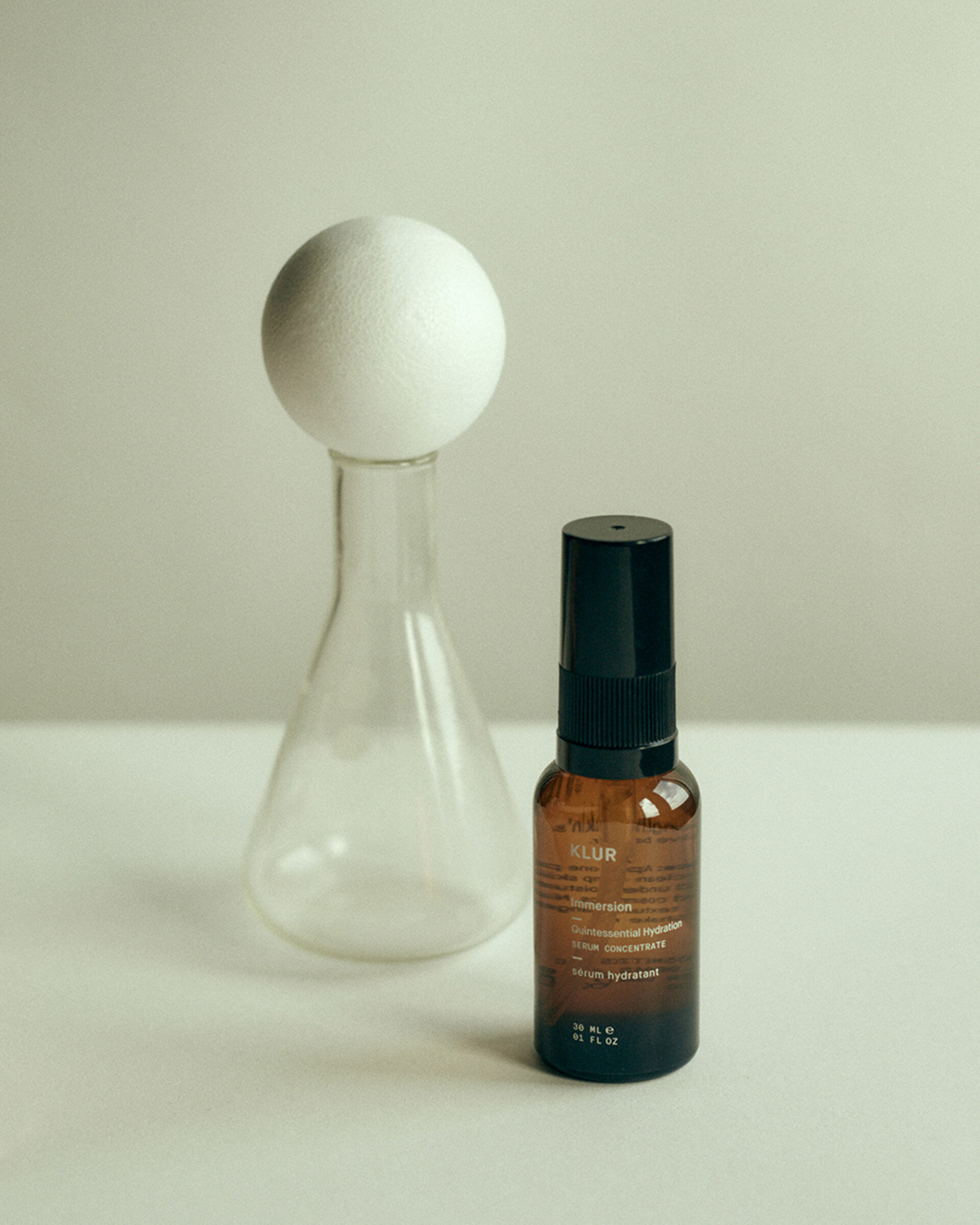 Amber glass bottle with black pump, situated near clear Erlenmeyer flask topped with white sphere on a white surface and neutral background.