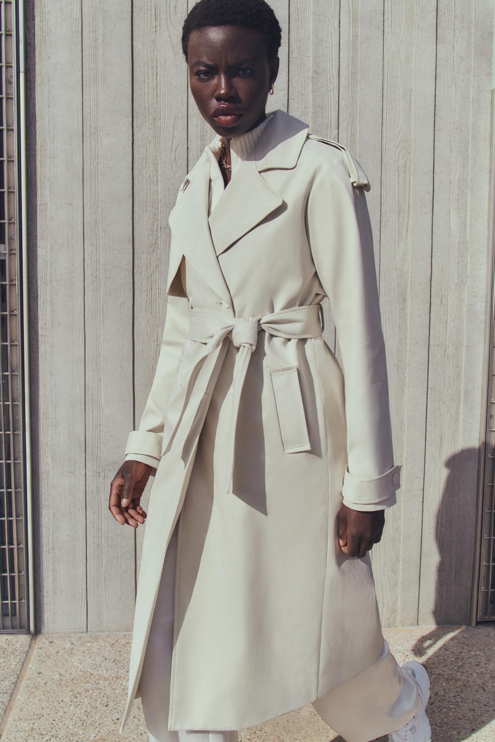 A Black woman stands in a beige belted trench coat on a concrete surface, with a light-colored wooden wall visible in the background.