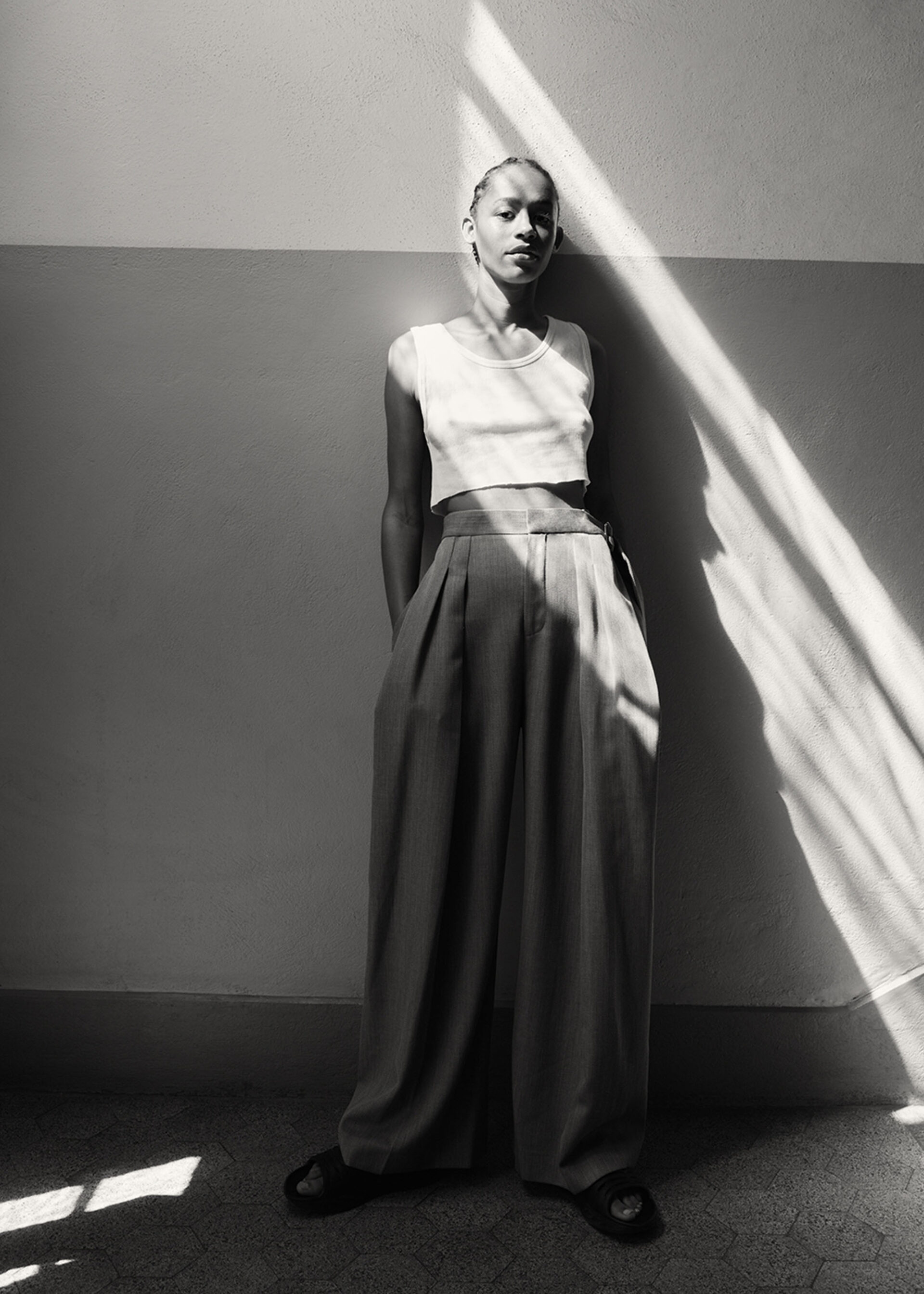 A stylish, young adult female poses in a cropped top and wide-legged trousers next to a sunlit wall in a black and white indoor shot.
