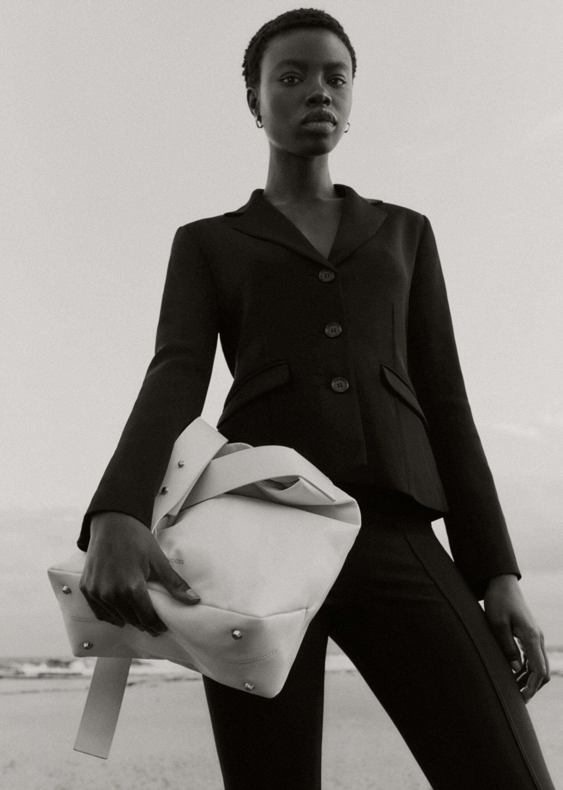 Black and white portrait of a Black woman in a blazer holding a large neutral bag at the beach with ocean waves behind her.
