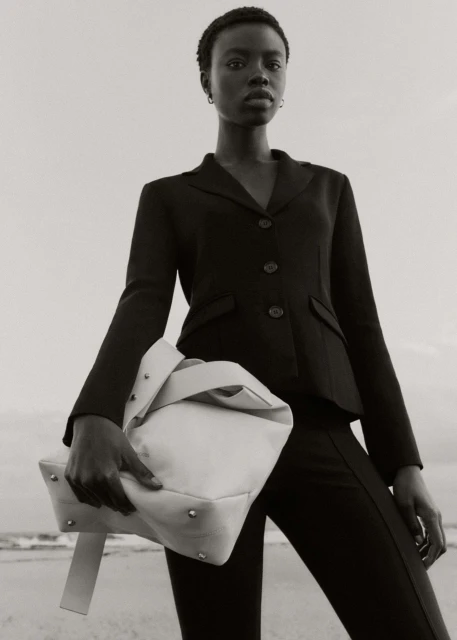 Black and white portrait of a Black woman in a blazer holding a large neutral bag at the beach with ocean waves behind her.