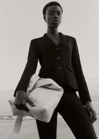 Black and white portrait of a Black woman in a blazer holding a large neutral bag at the beach with ocean waves behind her.