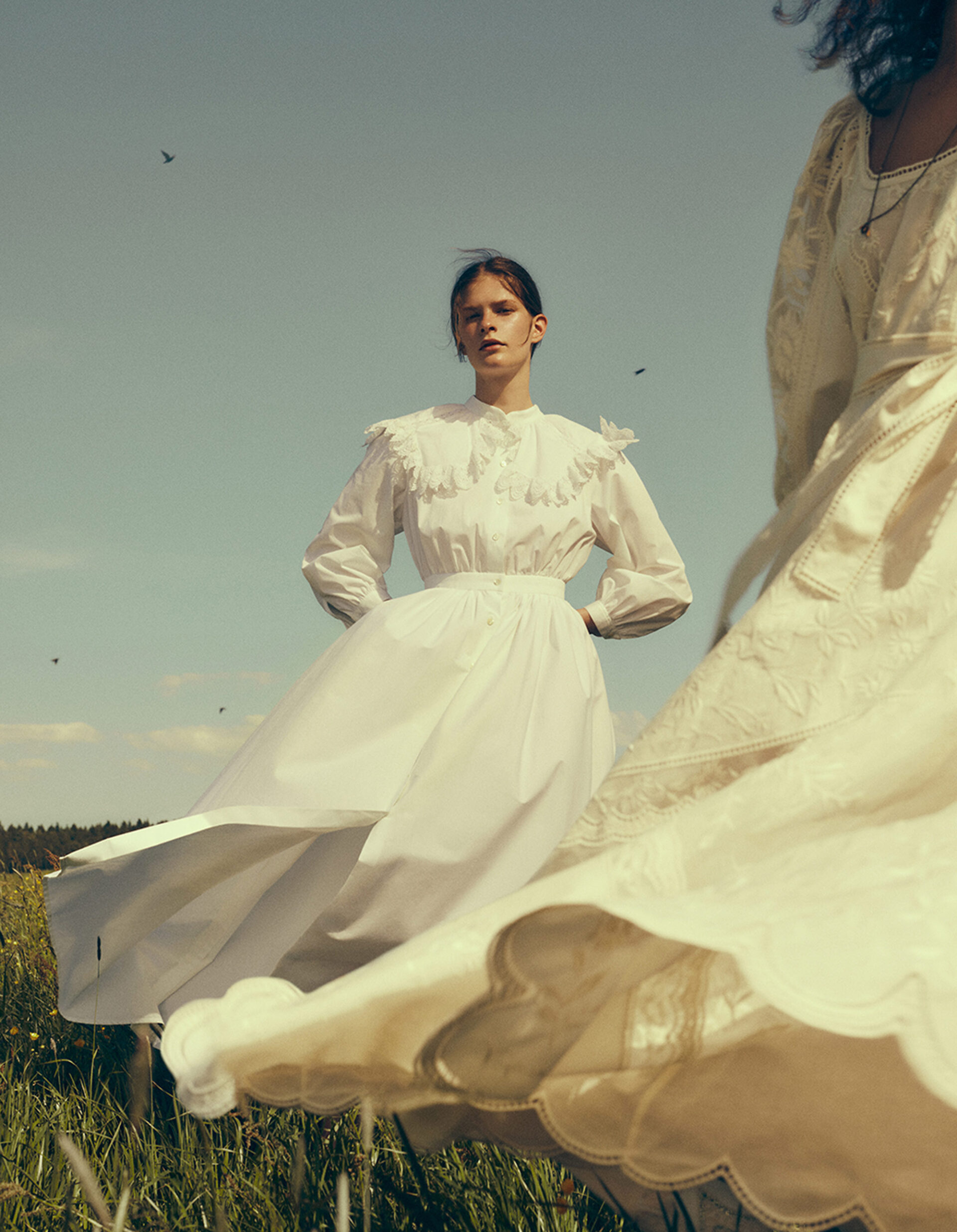 Two women in white dresses stand in a grassy field under a blue sky; one woman in focus, the other blurred and out of focus.