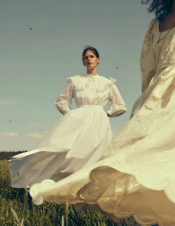 Two women in white dresses stand in a grassy field under a blue sky; one woman in focus, the other blurred and out of focus.