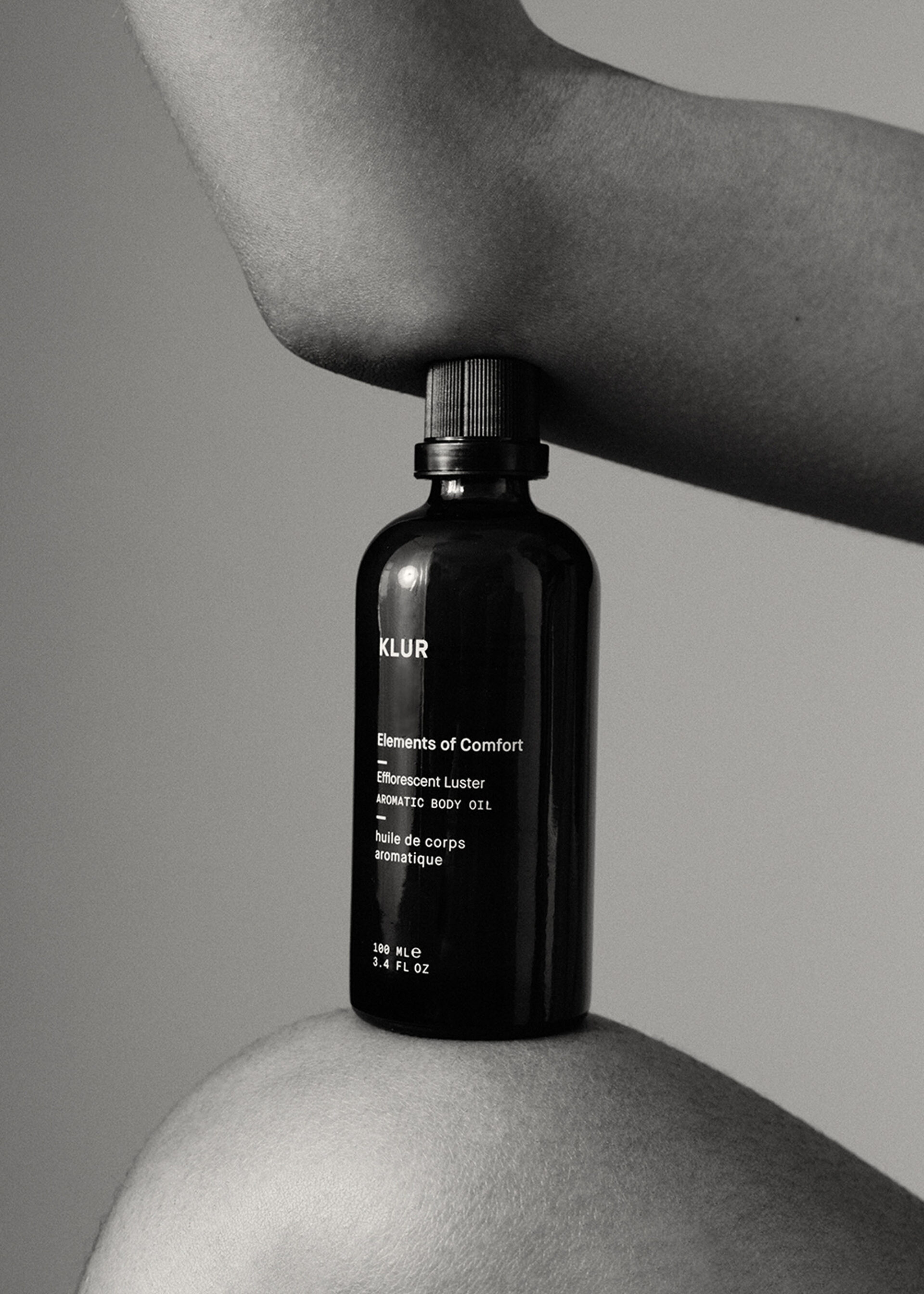 Monochromatic still life of KLUR Elements of Comfort aromatic body oil held between an adult's arm and shoulder on a plain background.
