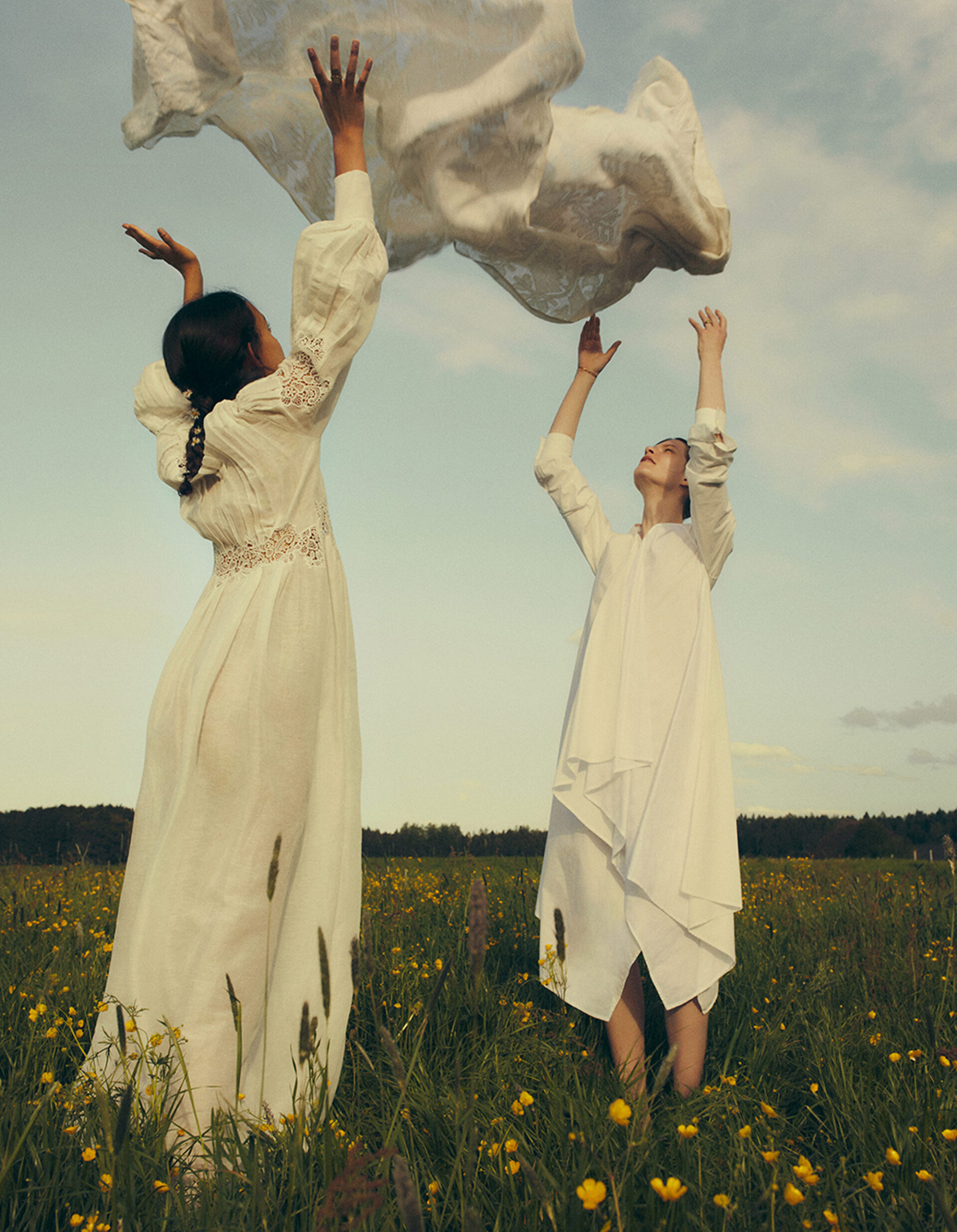 Two women in white dresses stand in a field of wildflowers with a sheer white cloth floating above them against a light blue sky.