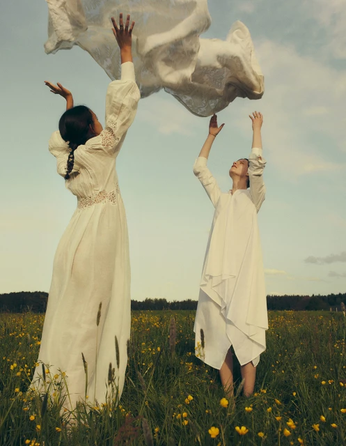 Two women in white dresses stand in a field of wildflowers with a sheer white cloth floating above them against a light blue sky.