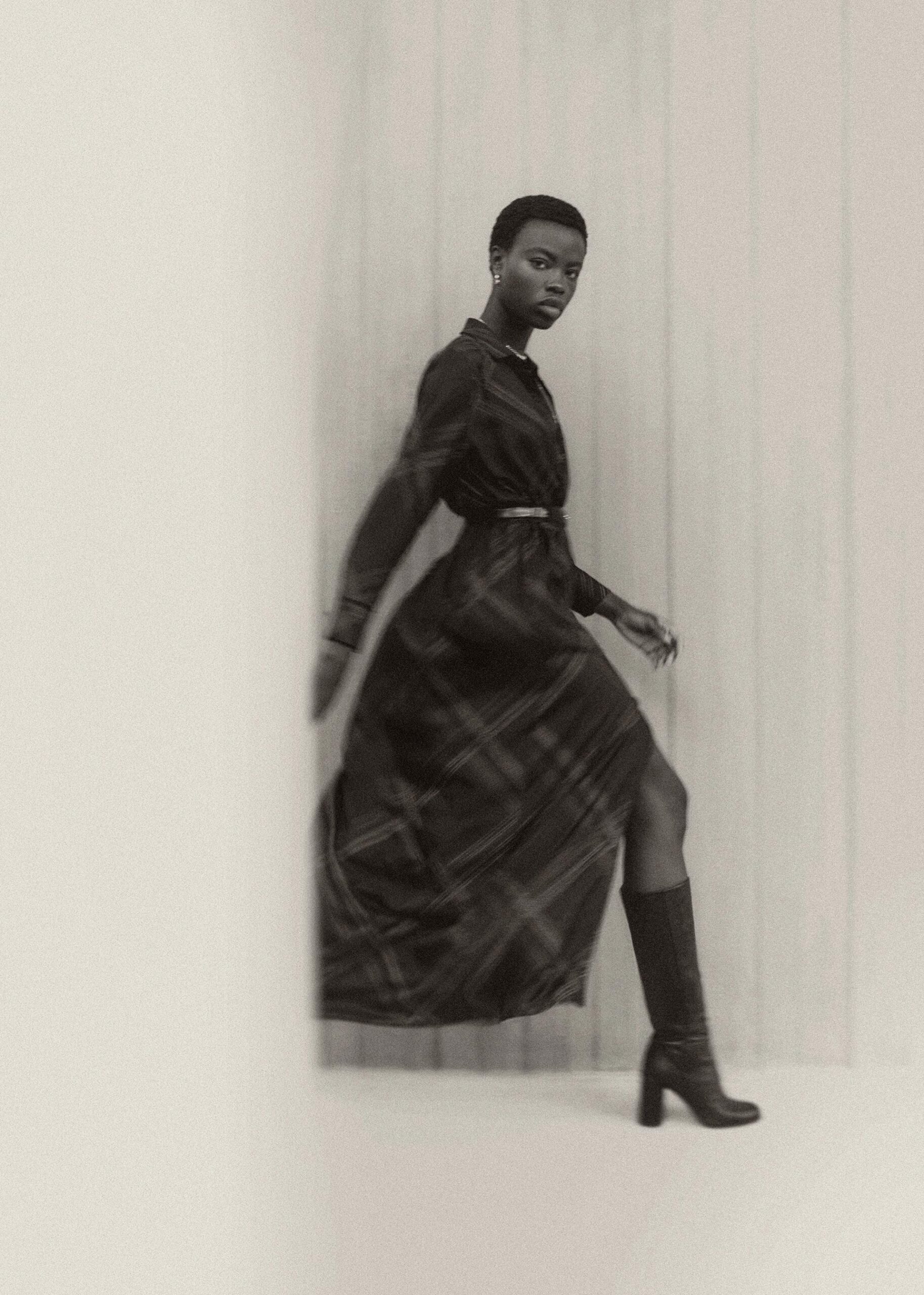 A Black woman in a plaid dress and boots walks in a minimalist studio setting, fashionably posed with diffused light.