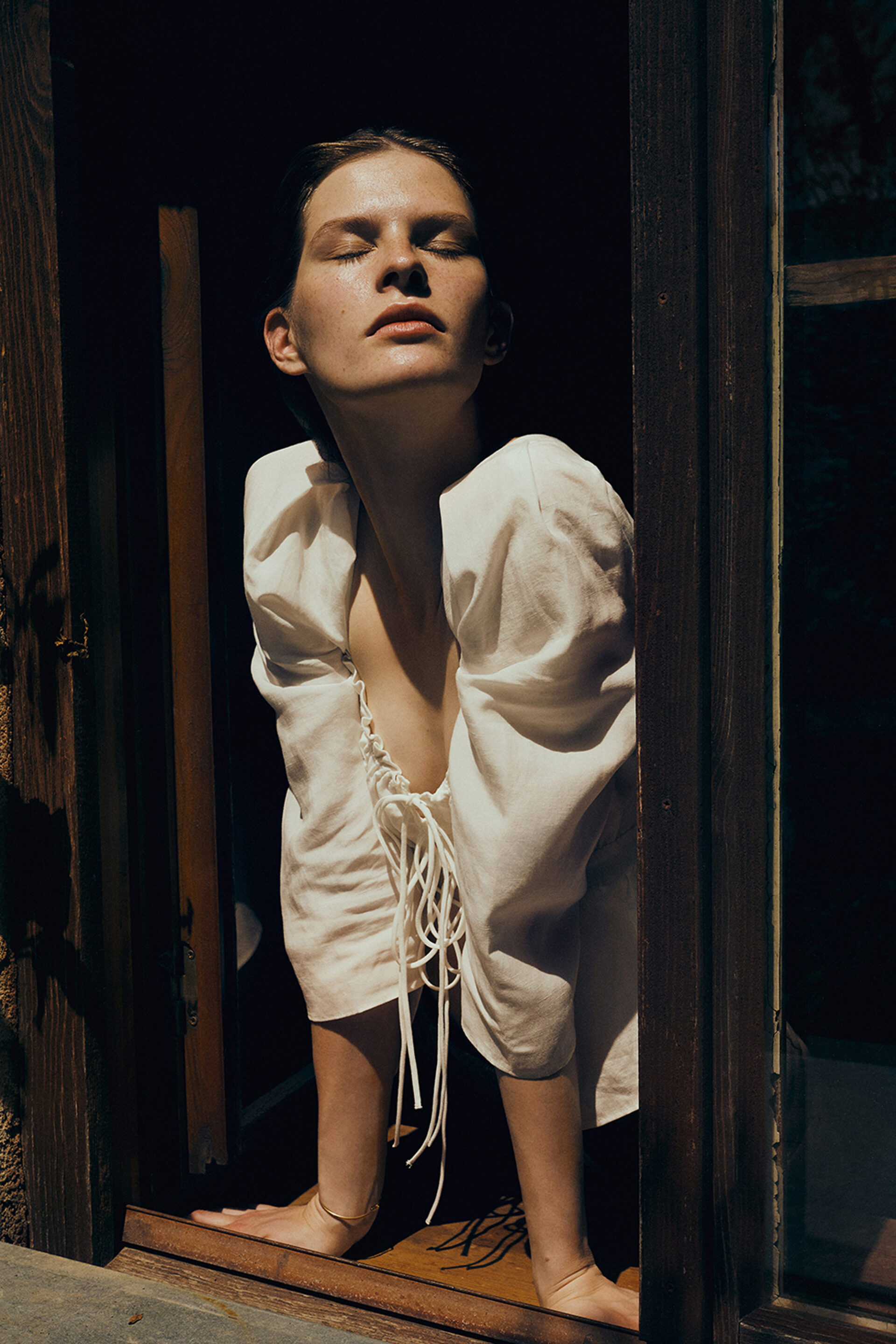 A light-skinned adult woman stands in a window frame wearing a white dress in a fashion setting on a sunny day.