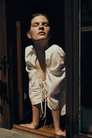 A light-skinned adult woman stands in a window frame wearing a white dress in a fashion setting on a sunny day.