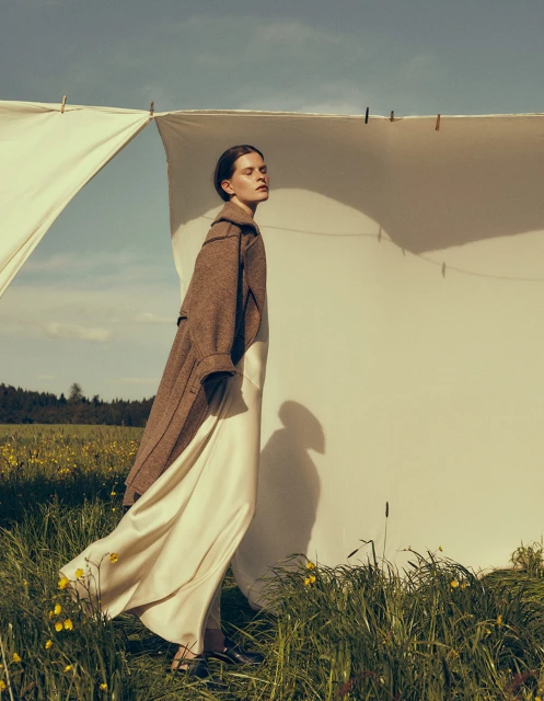 A woman in a long coat and dress stands outdoors in a grassy field near a white cloth backdrop on a clothesline under a blue sky.
