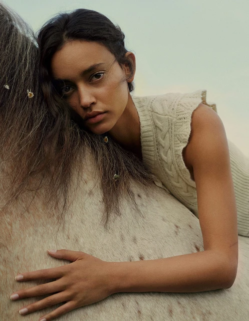 Woman rests on the back of a dappled grey horse outdoors, wearing a sleeveless cable knit sweater, with a dreamy, soft focus.