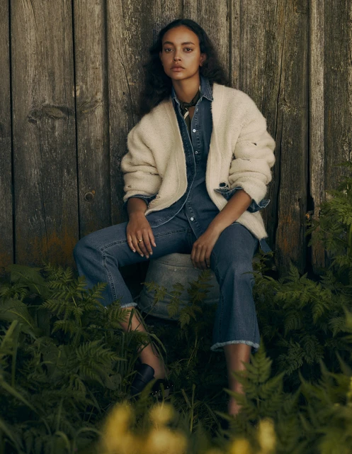 A woman sits on a bucket in front of a wood wall wearing a denim shirt, jeans, and a sweater in a rural outdoor setting.