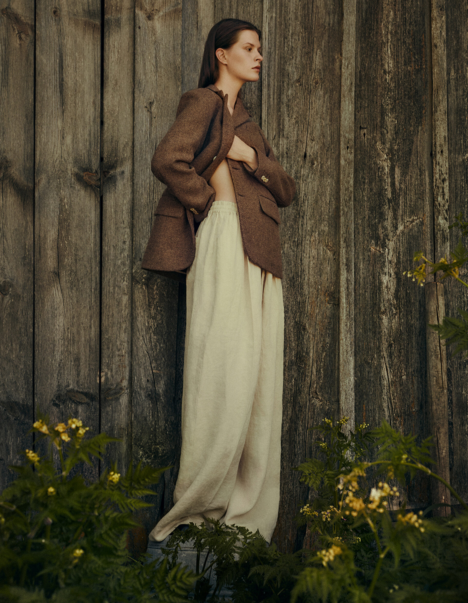 Woman poses in brown blazer and cream pants against weathered wooden wall with green foliage, captured in soft, natural lighting.