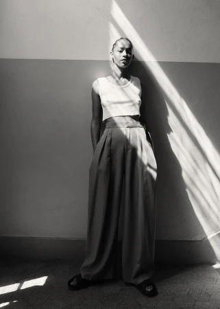 Monochrome portrait of a model standing against a wall with strong diagonal sunlight cutting across the scene.