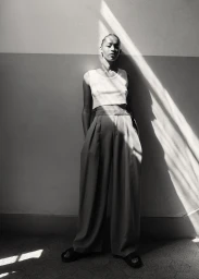 Monochrome portrait of a model standing against a wall with strong diagonal sunlight cutting across the scene.