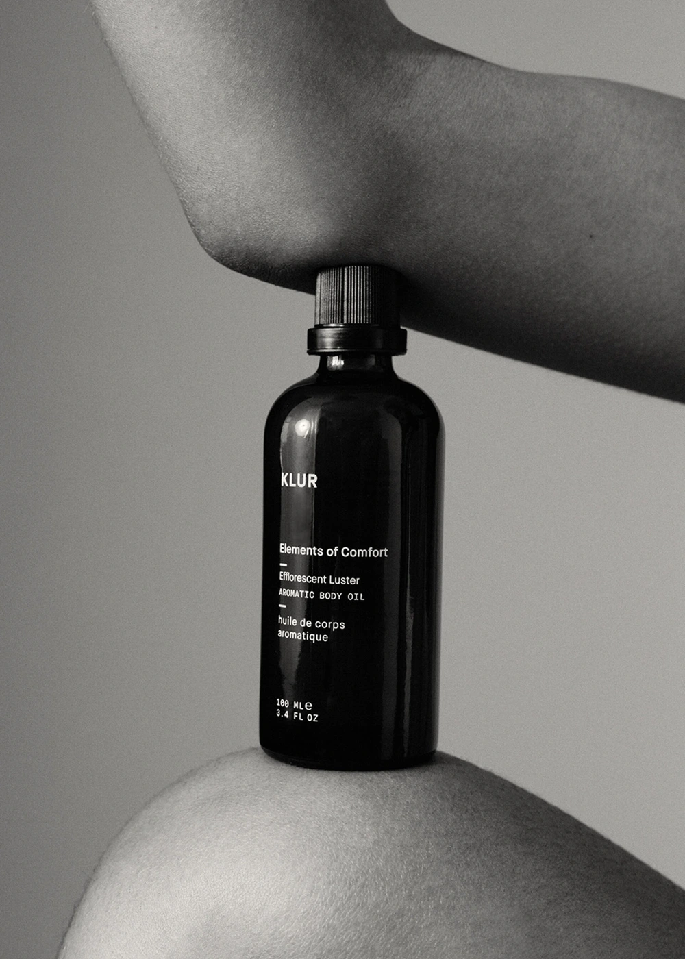 Black glass skincare bottle held horizontally between an elbow and a knee against a plain background