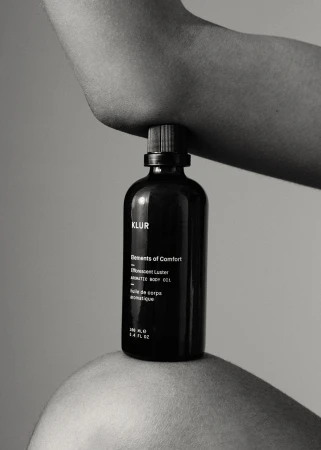 Black glass skincare bottle held horizontally between an elbow and a knee against a plain background