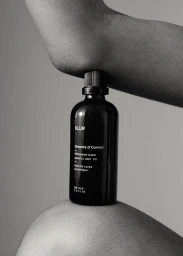 Black glass skincare bottle held horizontally between an elbow and a knee against a plain background