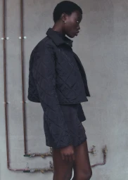 Black woman wearing a quilted black jacket and matching shorts against a concrete wall with exposed copper pipes