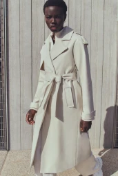 Black woman wearing a long white belted trench coat and white sneakers standing against a vertical paneled wall.