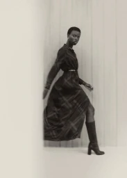 Black woman with short hair wearing a plaid long-sleeve dress and high boots posing against a wall.