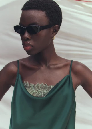 Black woman wearing dark sunglasses and a green satin camisole with lace trim against a pale background by Rahel Weiss.