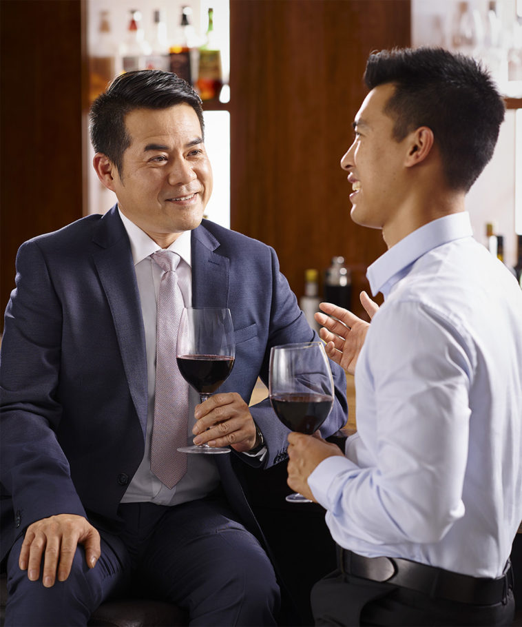 Two Asian businessmen in suits converse over glasses of red wine at an indoor bar, creating a sophisticated and engaging visual narrative.