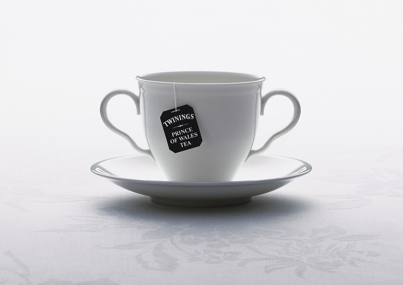 A white porcelain teacup and saucer are displayed against a soft white patterned background, with a tea bag tag attached to the cup.