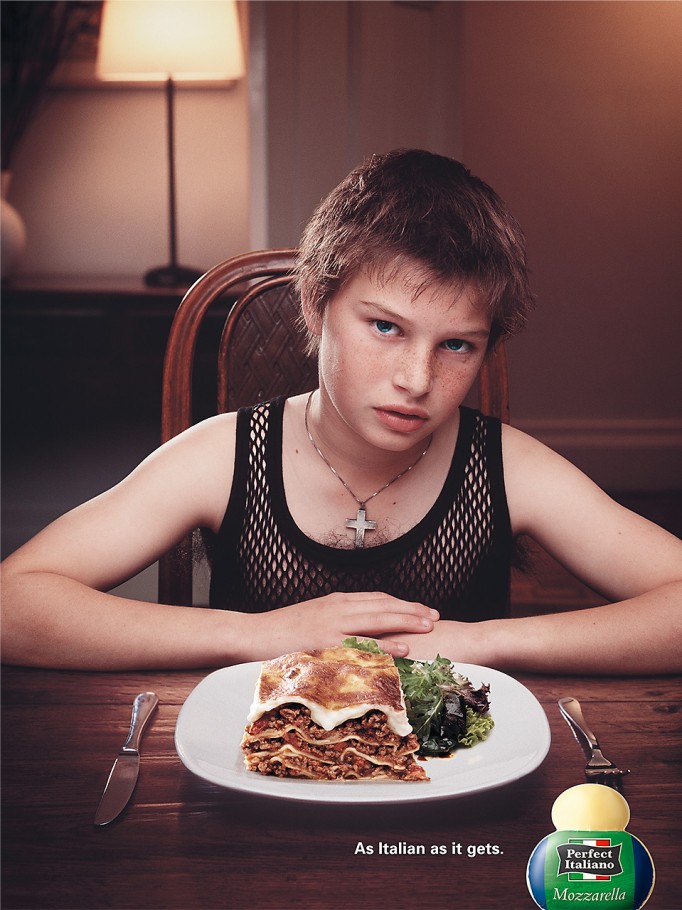 A preteen person sits at a table with lasagna and salad, knife and fork to either side. A bottle of Perfect Italiano mozzarella is visible.