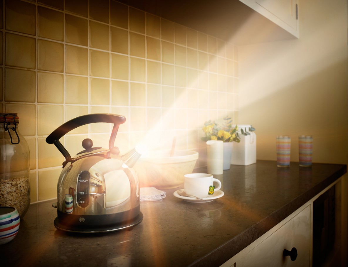 Shiny stainless steel kettle on a kitchen counter with tea cup, yellow flowers, and bright light streaming in for a cozy interior scene.